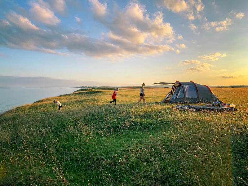 Picture showing a summer landscape in sunset. In the forground a tent a woman and two children camping