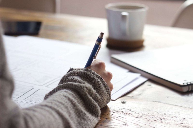 En kvinnas högra arm och hand, iklädd stickad ljusbrun tröjärm, skriver med en svart penna i ett skrivblock. I bakgrunden syns en vit kaffekopp med fat av trä på samma ljusbruna träbord.