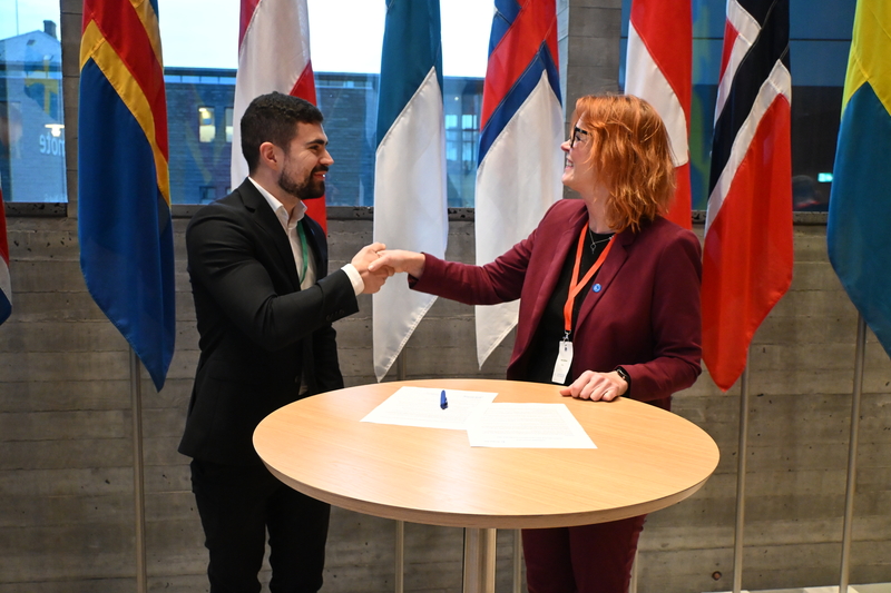Två personer undertecknar ett dokument framför de nordiska flaggorna
