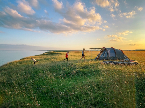 Picture showing a summer landscape in sunset. In the forground a tent a woman and two children camping